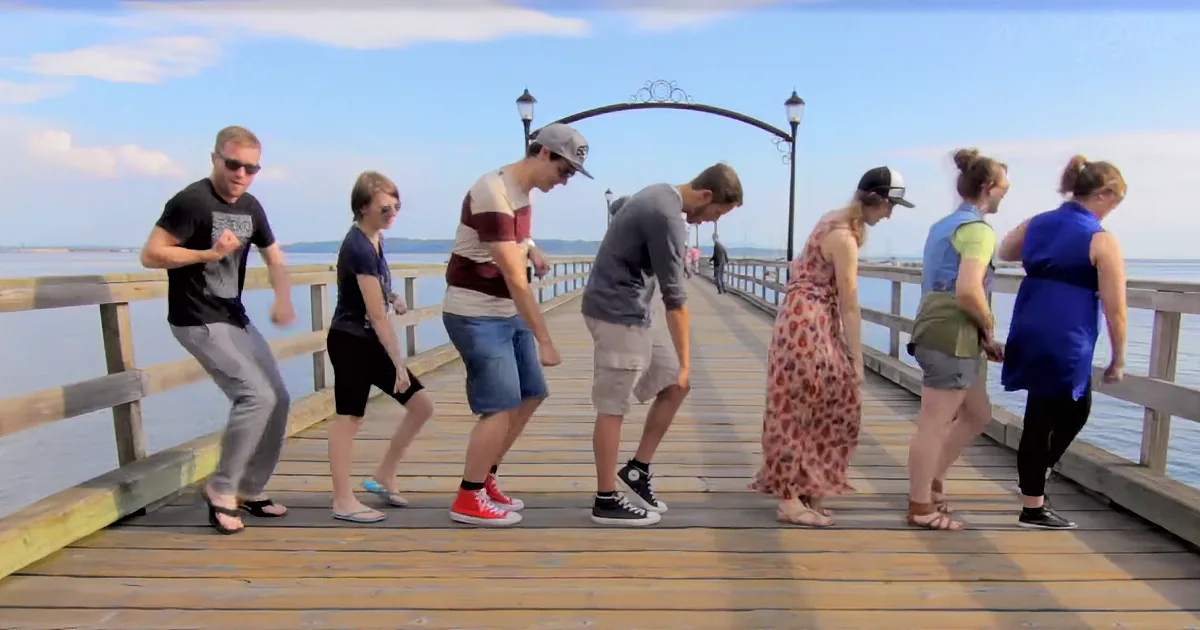 A group of six people dancing on a wooden pier by the water under a clear sky.