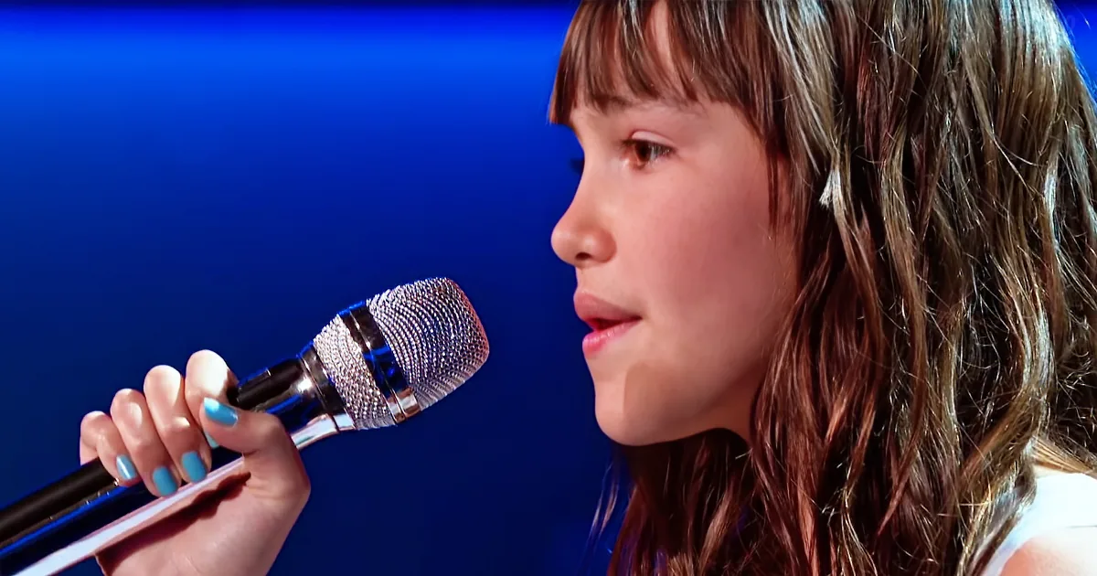A young girl with long brown hair holds a microphone, her nails painted blue, against a blue background.