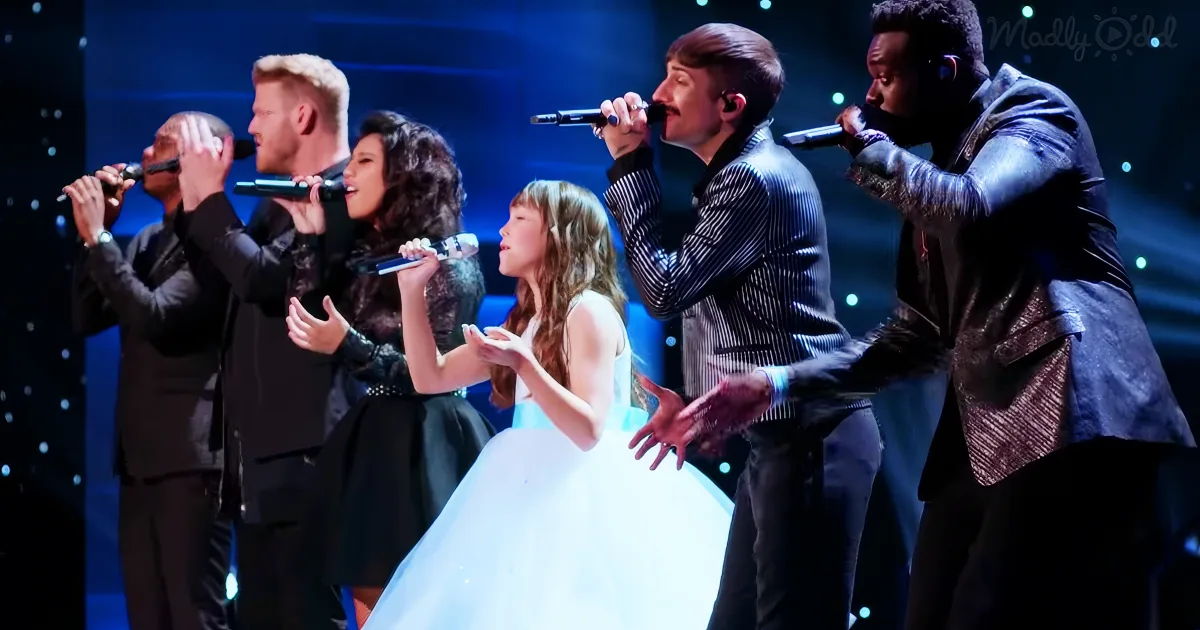 A young girl in a white dress sings surrounded by five male singers on stage.