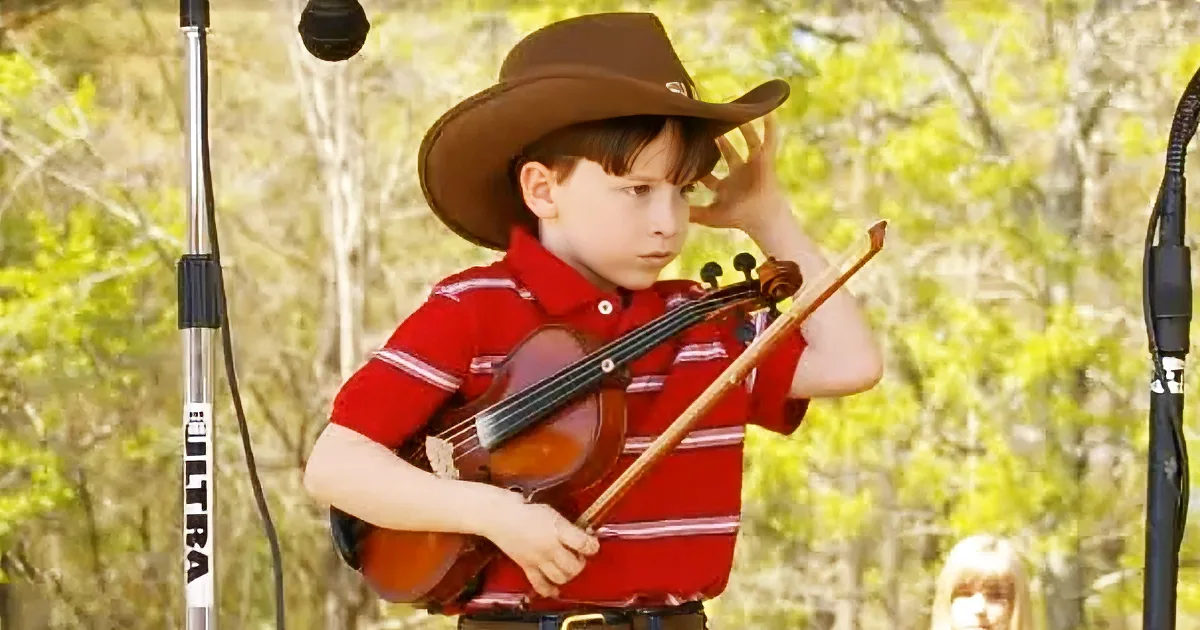 A young boy in a red shirt and cowboy hat plays a violin, surrounded by trees and microphones.