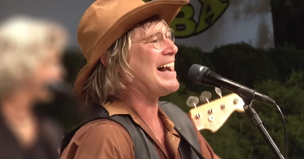 A person wearing a brown hat and glasses sings into a microphone, with a guitar and another person in the background.