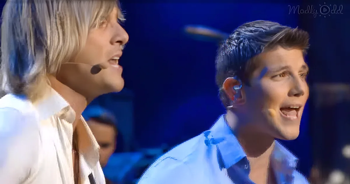 Two individuals, one with long blonde hair and the other with short dark hair, are singing on stage.