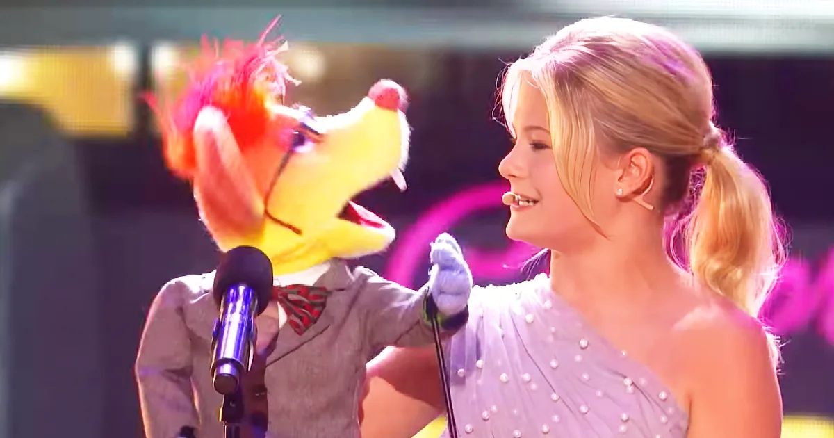 A woman in a pearl dress interacts with a puppet in a suit, both holding microphones.