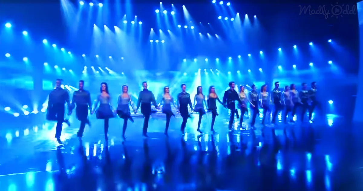 A group of dancers in traditional attire holds hands on a stage illuminated by blue lights.