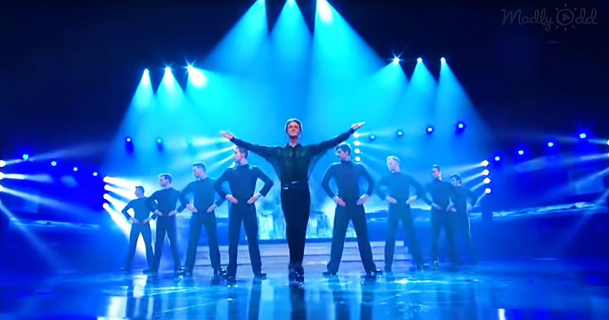 A man in a black outfit stands center stage with arms outstretched, surrounded by dancers in matching attire under blue...
