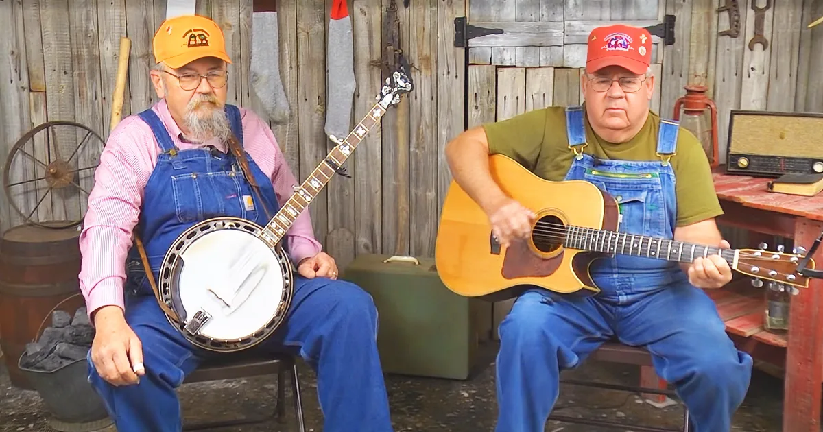 Two men in overalls play banjo and guitar in a rustic setting with wooden walls and tools.