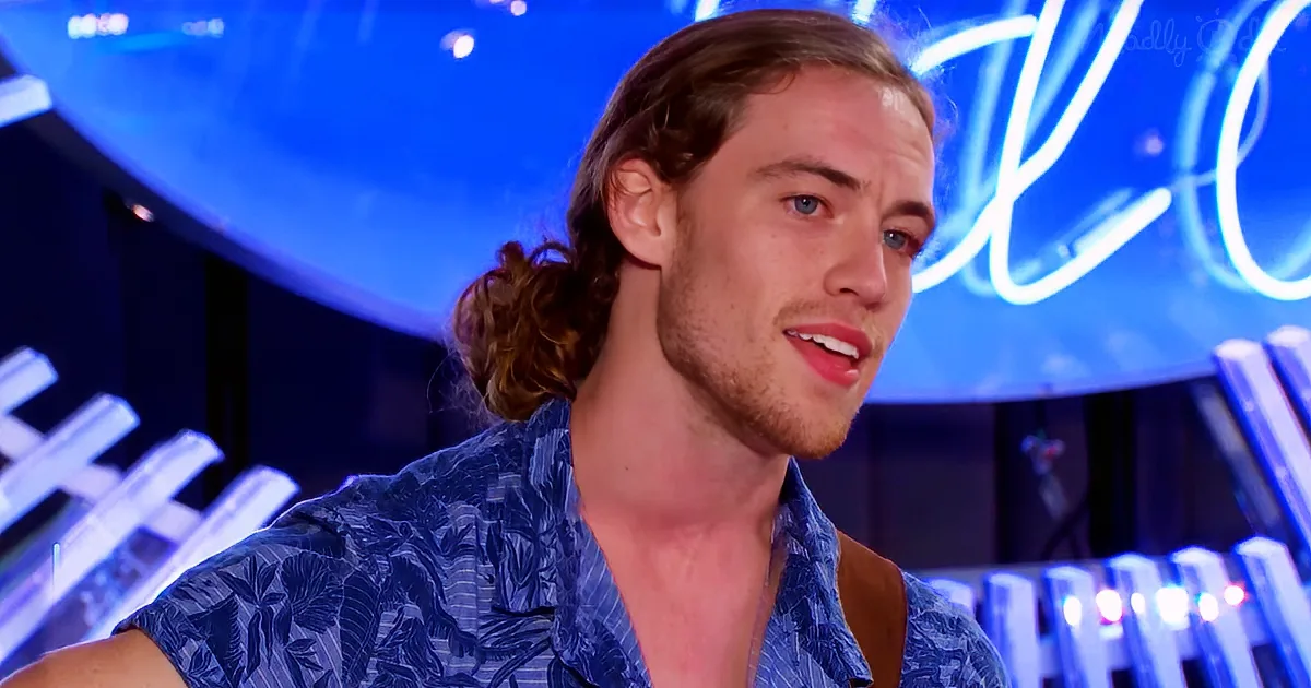 A man with long hair in a ponytail, wearing a blue floral shirt, is speaking in front of a blue-lit background.