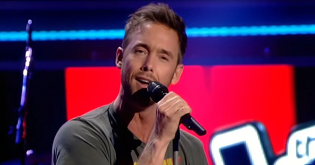 A man with short hair holds a microphone, singing on a stage with red and blue lighting.