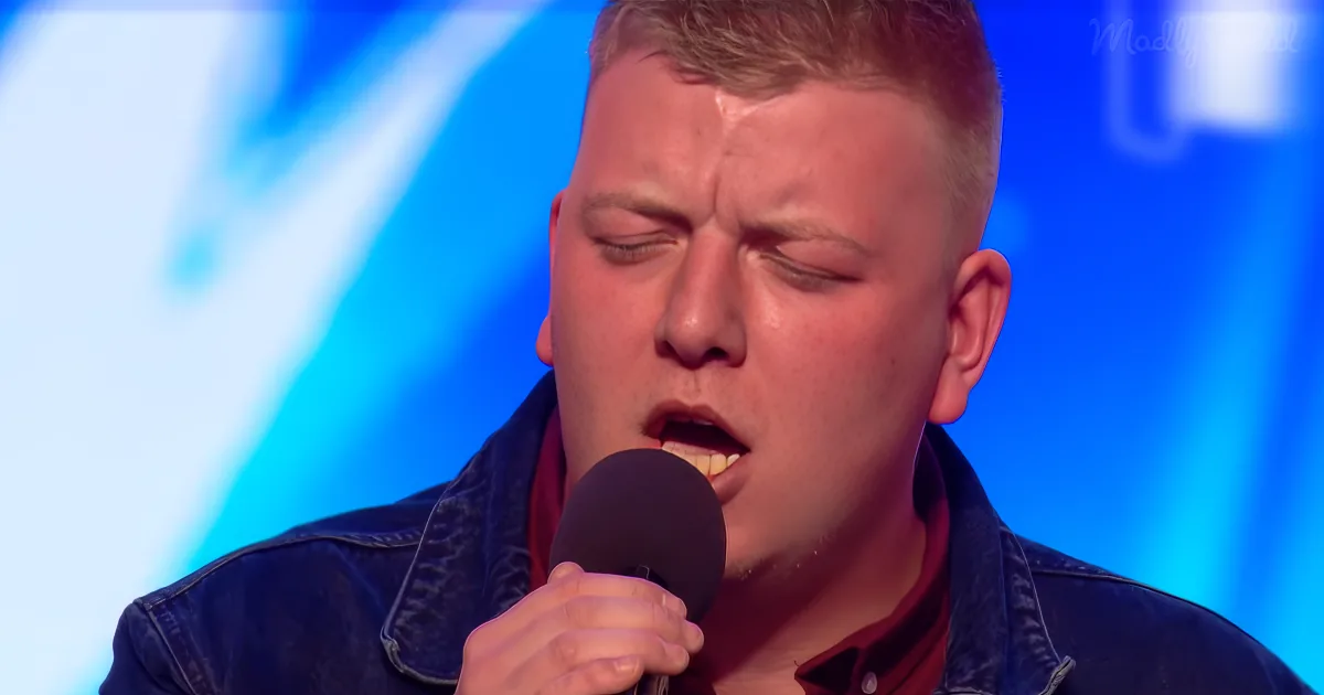 A man in a denim jacket sings into a microphone, eyes closed, against a blue background.