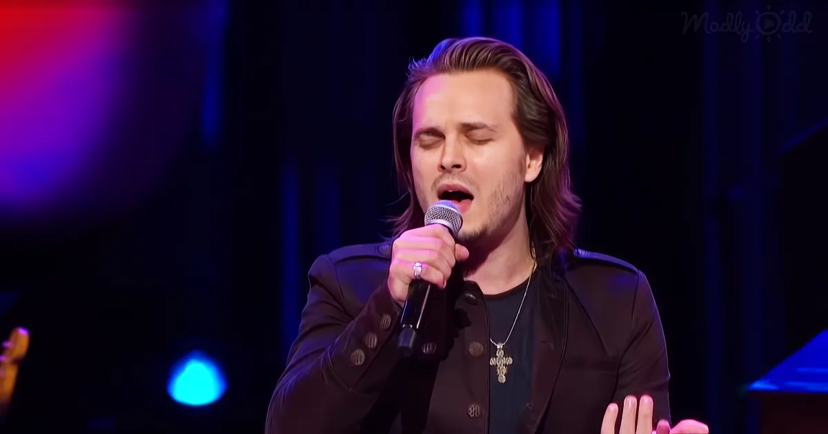 A man with long hair and a cross necklace sings into a microphone on stage.