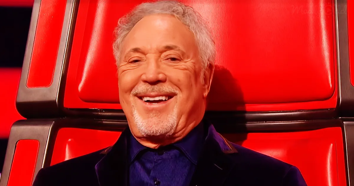 A smiling older man with gray hair and a beard, wearing a dark suit, sits in a red chair.