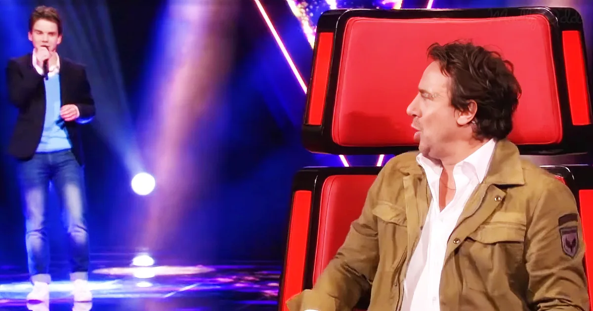 A man in a brown jacket sits on a red chair, watching another man in a blue shirt perform on stage.