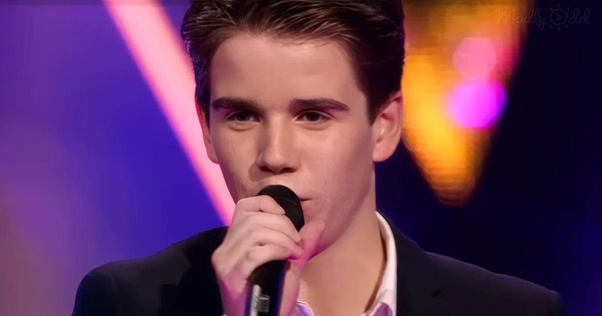 A young man in a suit holds a microphone, singing on stage with colorful lights behind him.