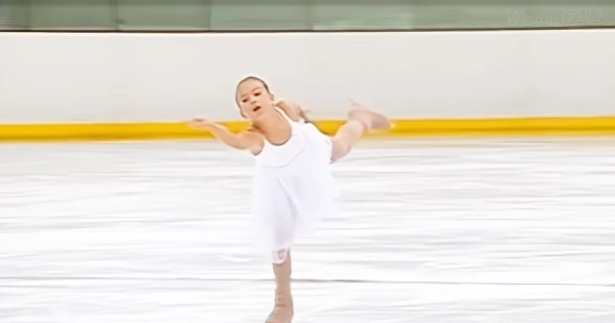 A young girl in a white dress skates gracefully on an ice rink, arms outstretched.