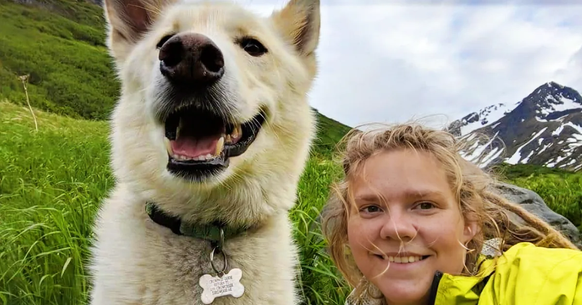 A happy white dog with a green collar smiles alongside a person in a yellow jacket against a backdrop of green grass and...