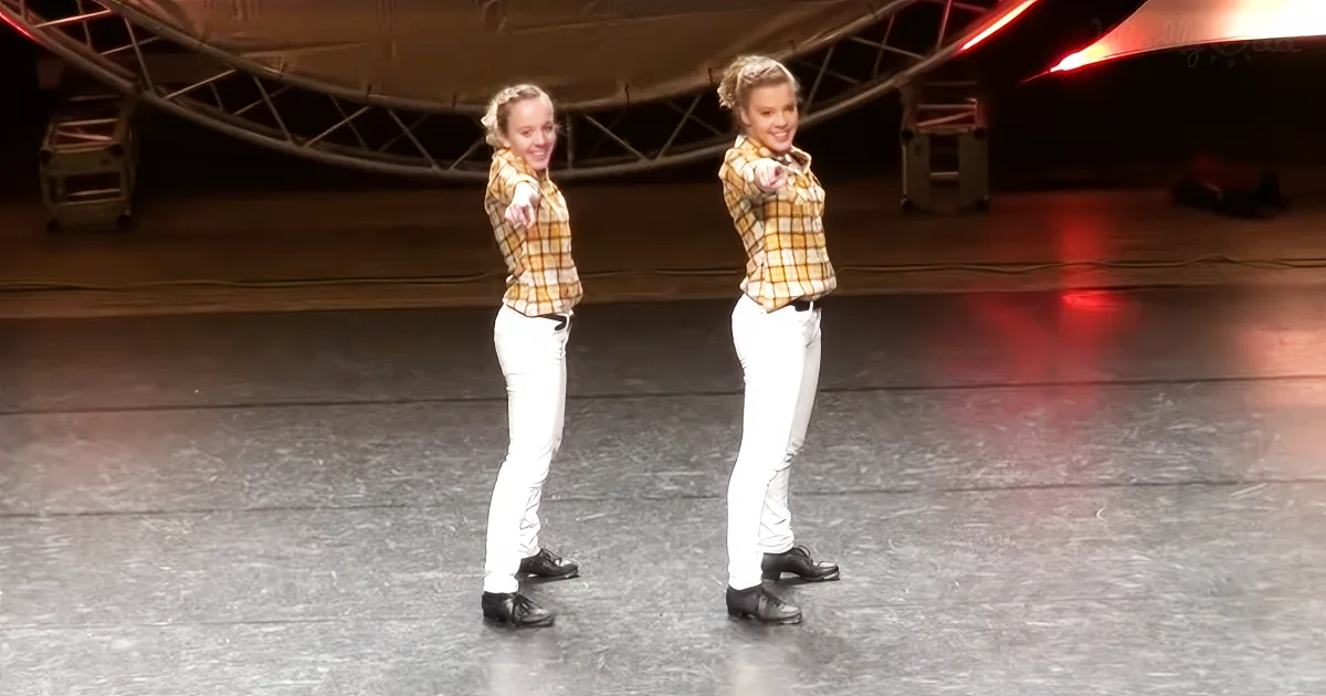 Two identical figures in plaid shirts and white pants stand on a stage, pointing forward.
