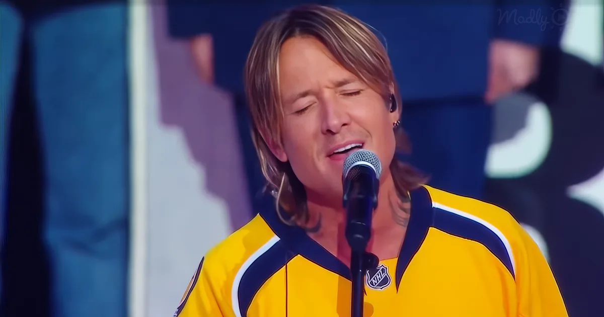 Keith Urban sings into a microphone, wearing a yellow jersey with a NHL logo, in a performance setting.