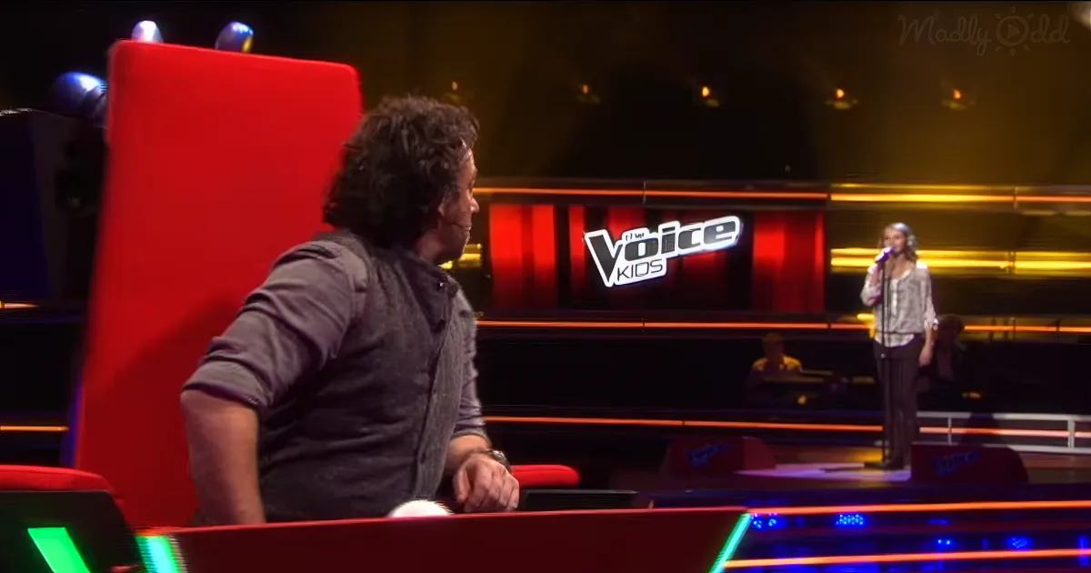 A man sits in a red chair on a stage, facing a young singer on a microphone stand.