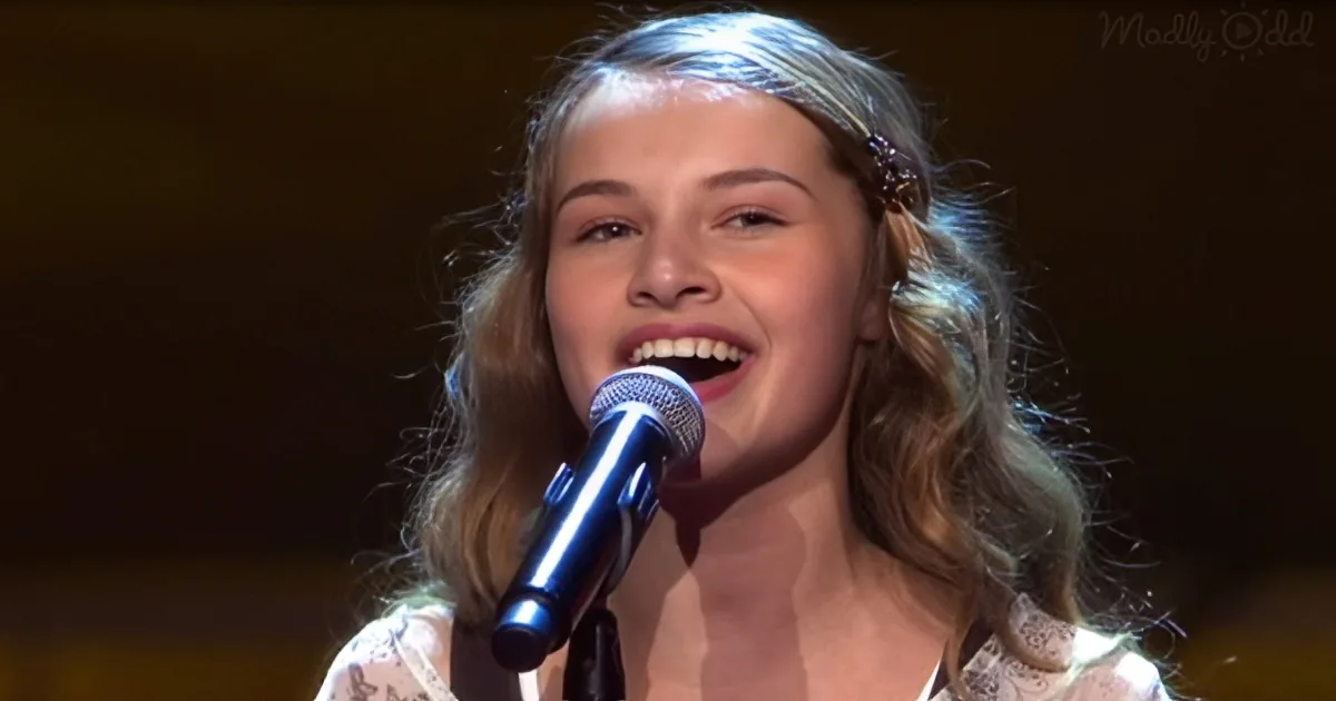 A woman with long, wavy hair sings into a microphone, her mouth open, wearing a white top.