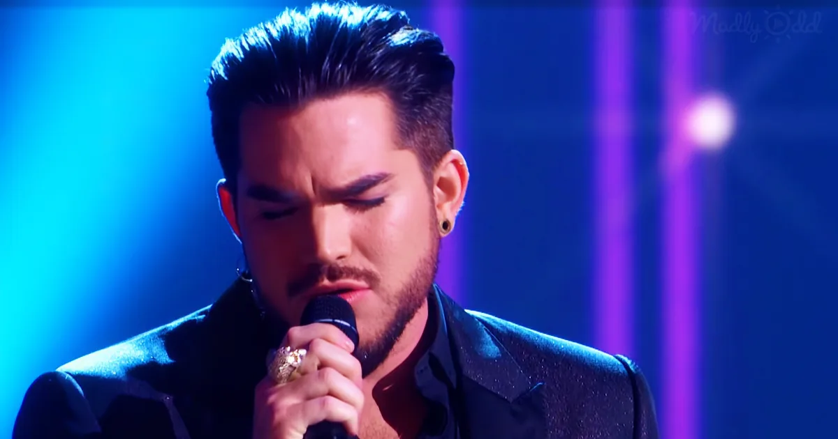 A man with dark hair and beard, wearing a black suit, sings into a microphone under blue and purple stage lights.