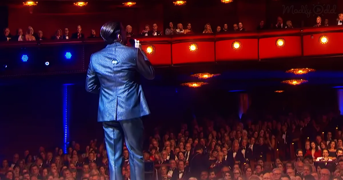 A person in a blue suit stands on stage, facing an audience in a theater with red and blue lighting.