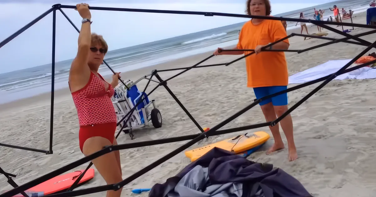 A woman in a red swimsuit is adjusting a beach canopy on the sand, with another person in an orange shirt assisting nearby.