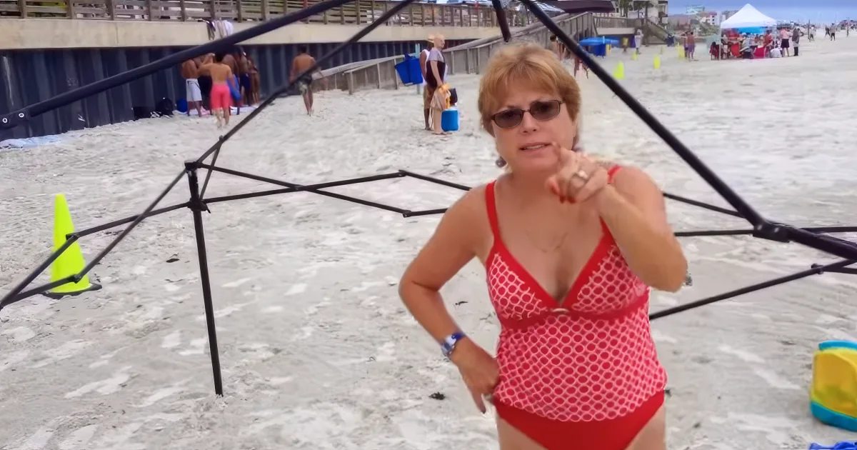 A woman in a red swimsuit stands on a beach, pointing forward, with a black umbrella and yellow cone nearby.