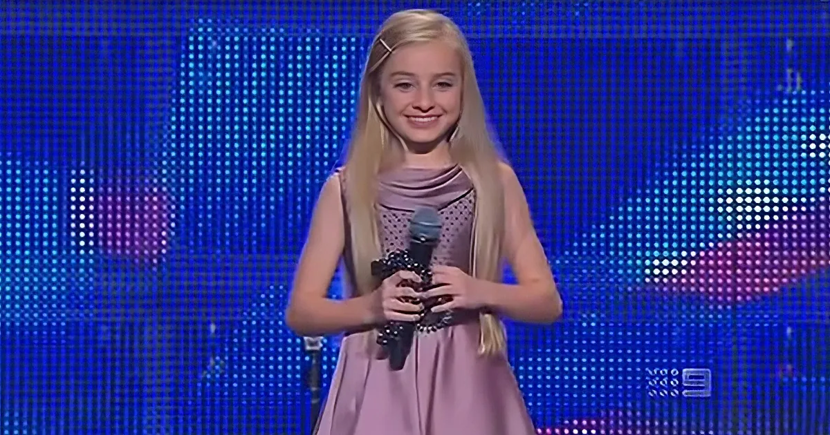 A young girl with long blonde hair, wearing a pink dress, holds a microphone on stage with a blue LED background.