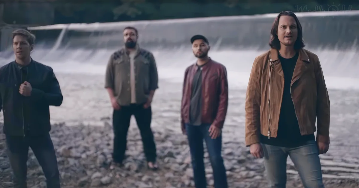 Four men stand on a rocky shore, dressed casually, with a dam and flowing water in the background.