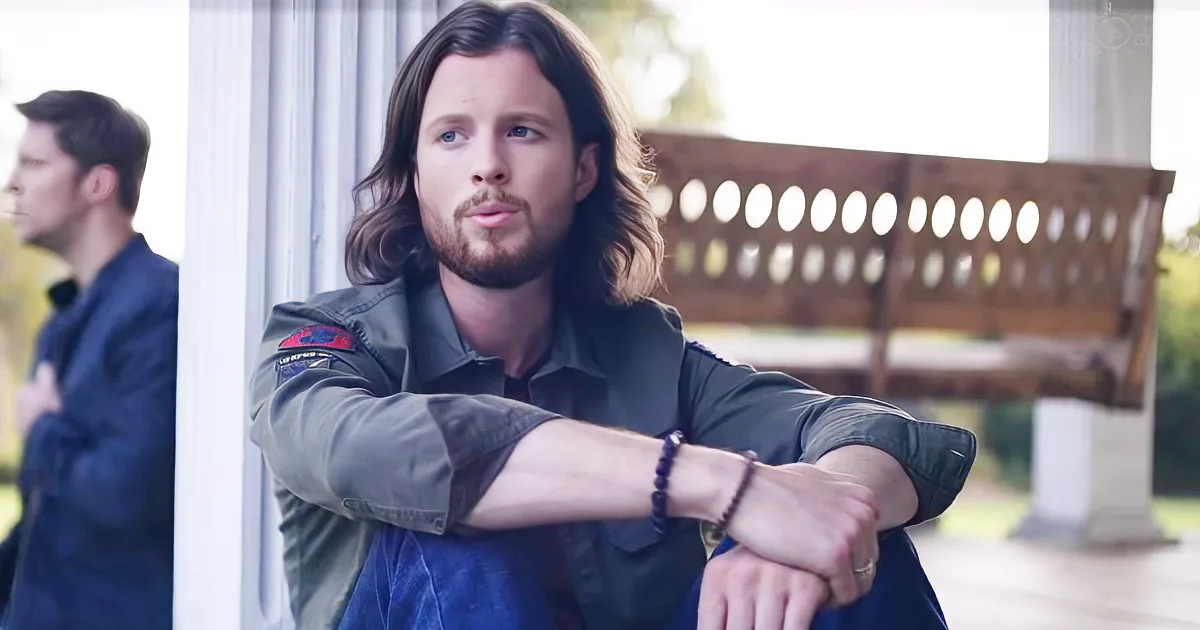 Man with long hair and beard, wearing a green jacket, sits with arms crossed, looking to the side.