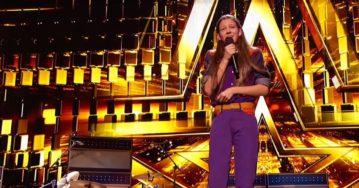 A young girl in purple pants and a purple shirt sings into a microphone on a stage with a golden geometric background.