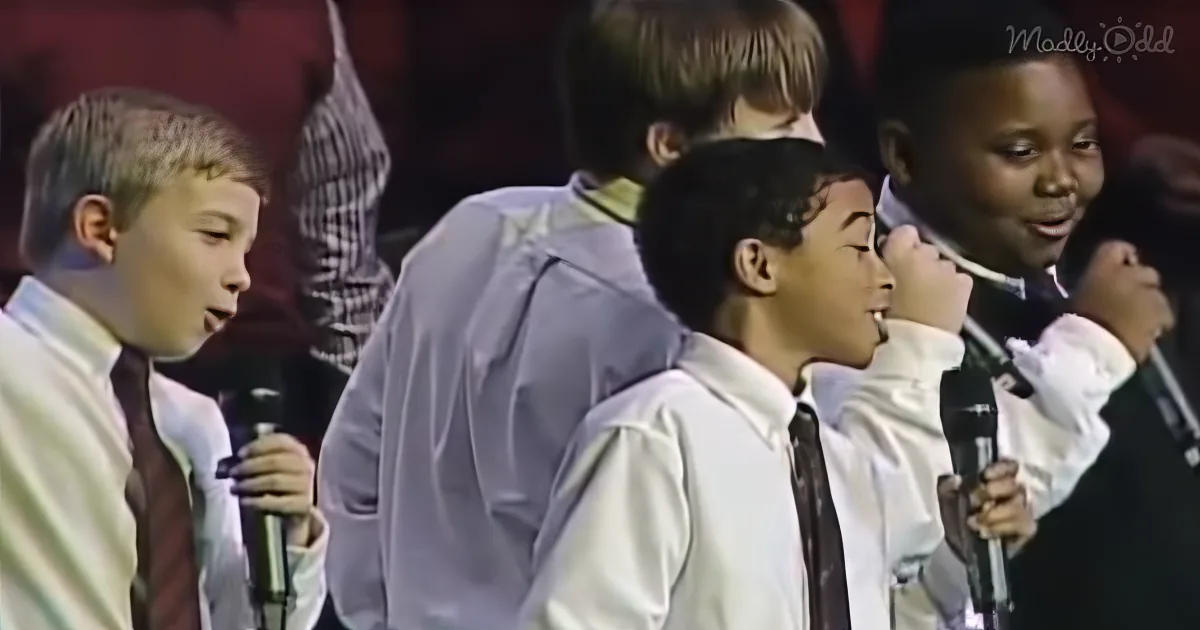 Four boys in white shirts and ties sing into microphones, one boy in the middle leans forward.