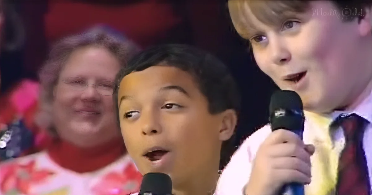 A young boy sings into a microphone, holding it with his right hand, while another child looks on.