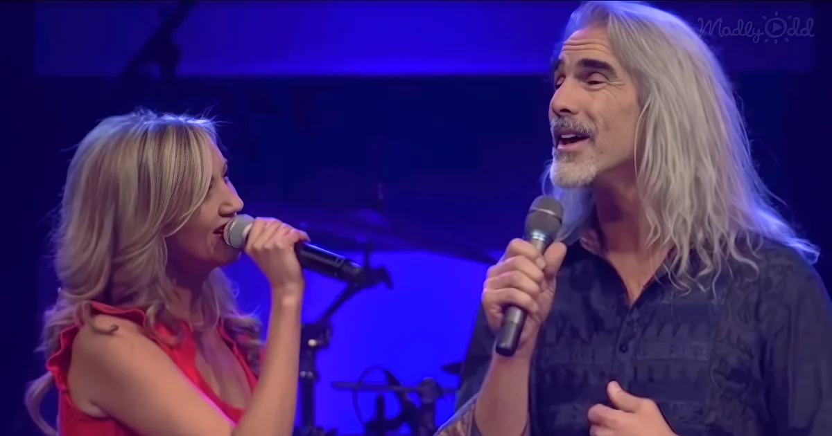A man and woman sing into microphones on stage under blue lighting.