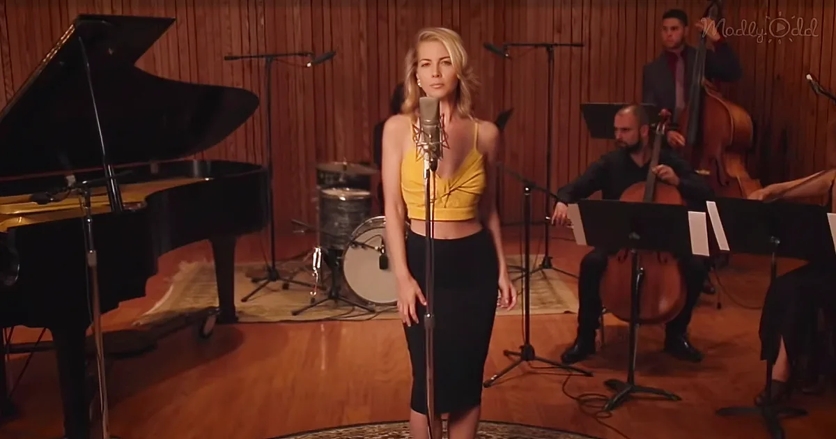 A woman in a yellow top and black skirt sings into a microphone, surrounded by musicians and instruments.