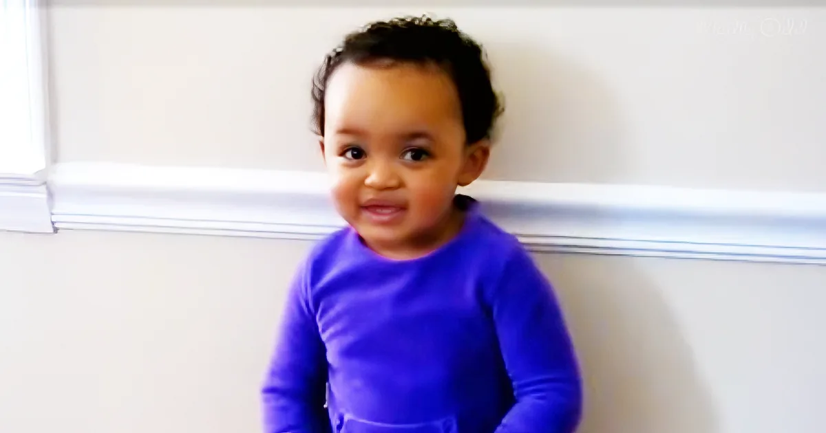 A toddler in a purple shirt sits against a white wall, looking slightly to the side.