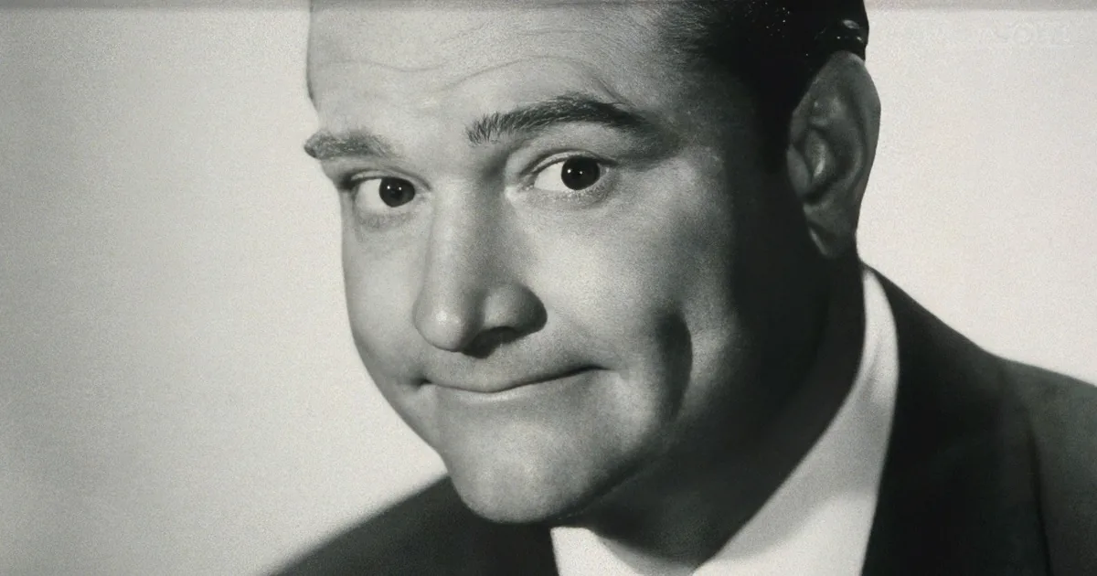 A man in a suit and tie, smiling with a surprised expression, in a black-and-white portrait.
