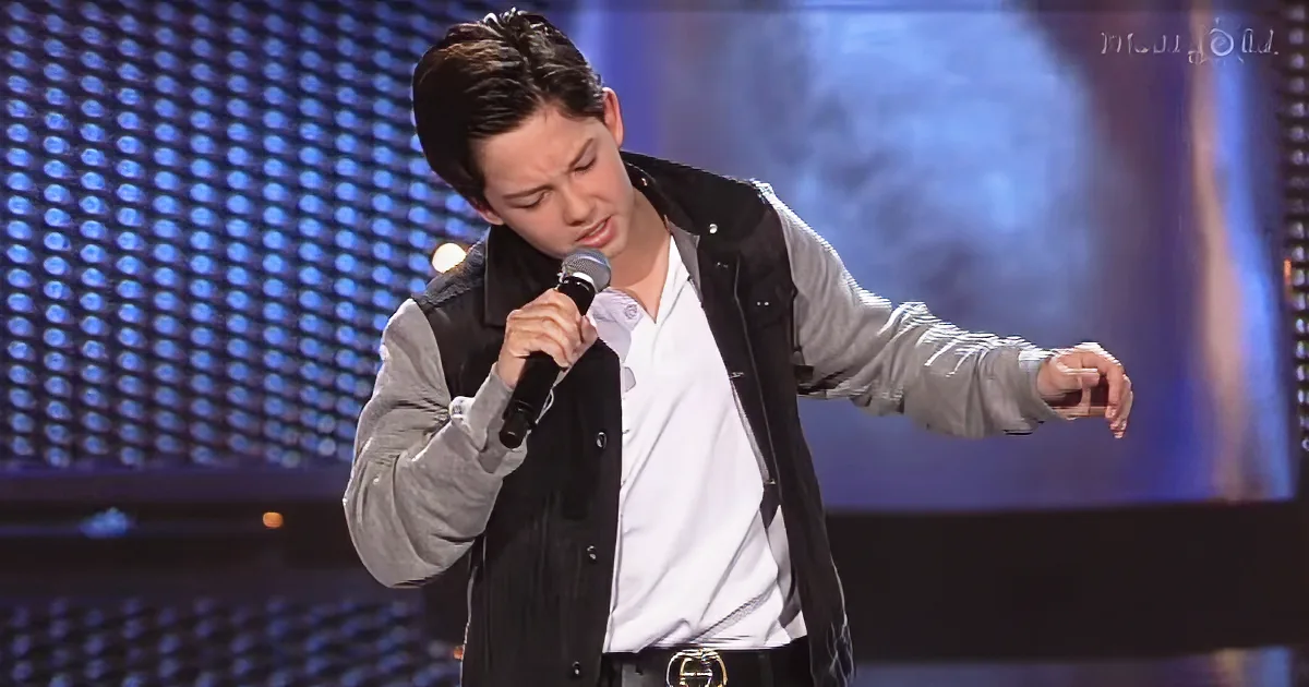 A man in a black vest and white shirt sings into a microphone on stage.