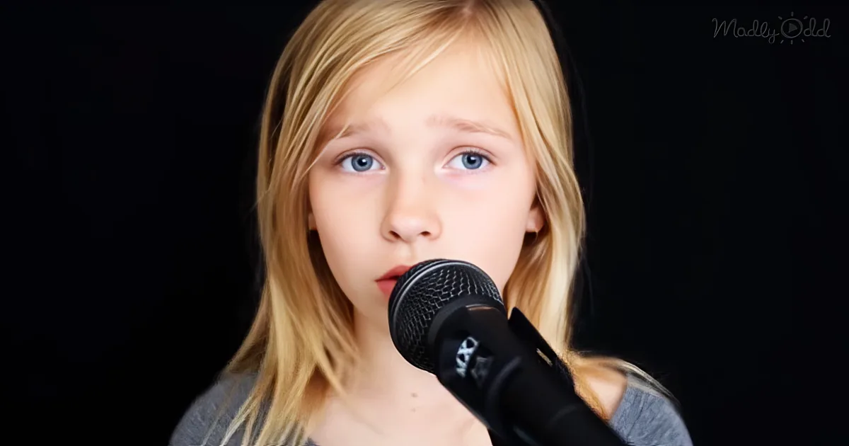 A young girl with blonde hair and blue eyes sings into a microphone against a dark background.