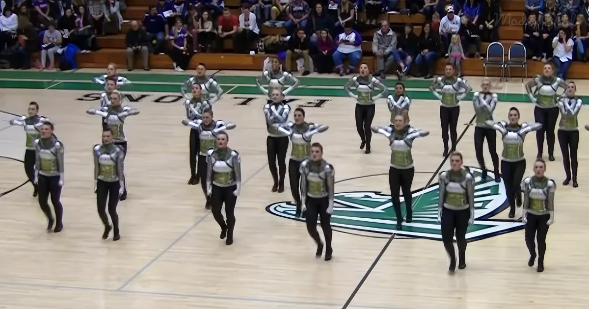 A synchronized dance team in silver and green uniforms performs on a basketball court.