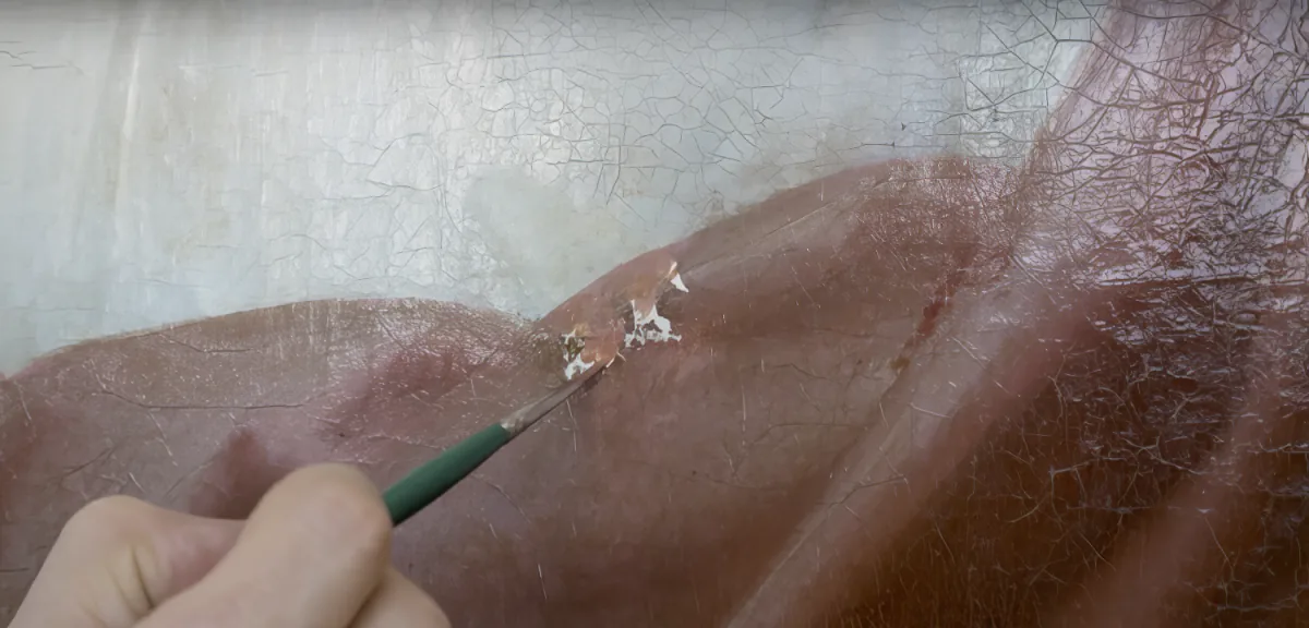 A hand holds a small tool, carefully working on a textured surface with visible cracks and a reddish-brown hue.
