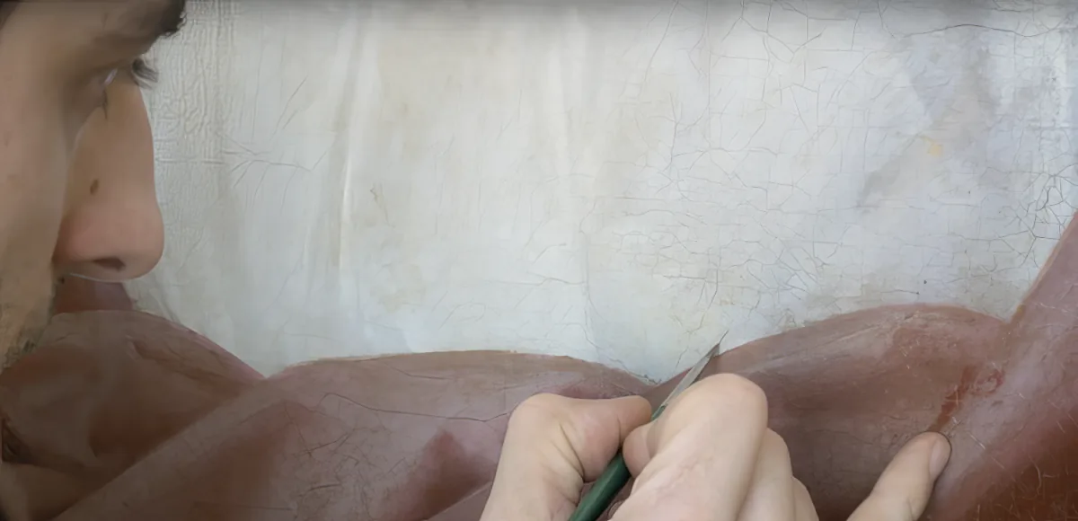 A person is carefully painting or drawing on a textured surface with a green tool.
