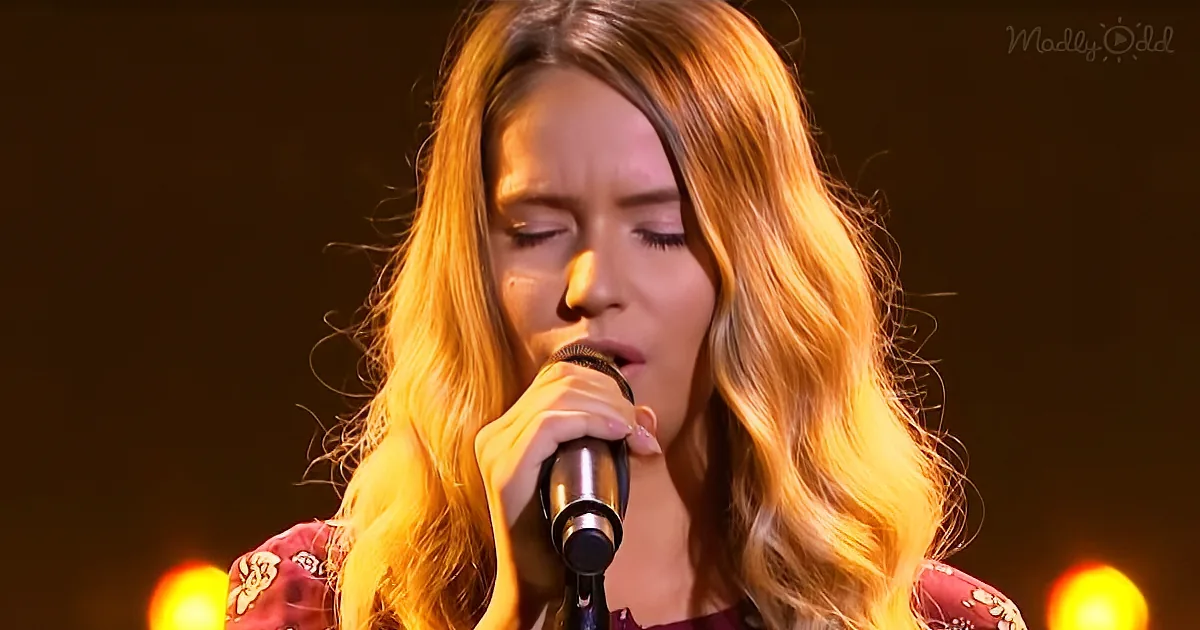 A person with long, wavy blonde hair sings into a microphone, wearing a patterned top, with warm stage lighting in the...