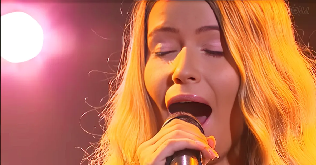 A woman with long, wavy hair sings into a microphone, illuminated by stage lights.