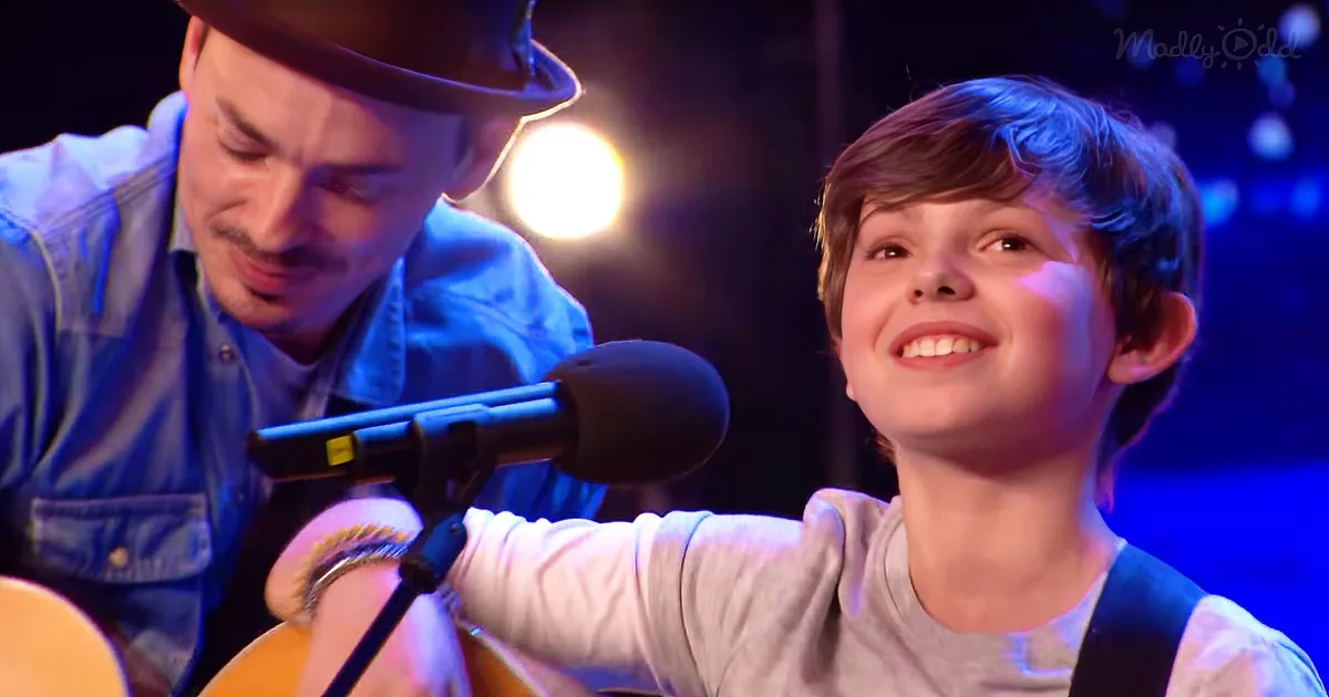A young boy smiles while playing guitar, with a man in a hat nearby, both under stage lights.