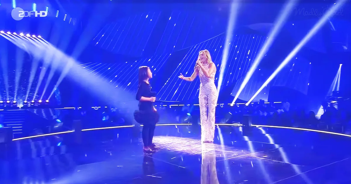 A woman in a sparkly dress sings on stage with another woman, illuminated by blue lights.