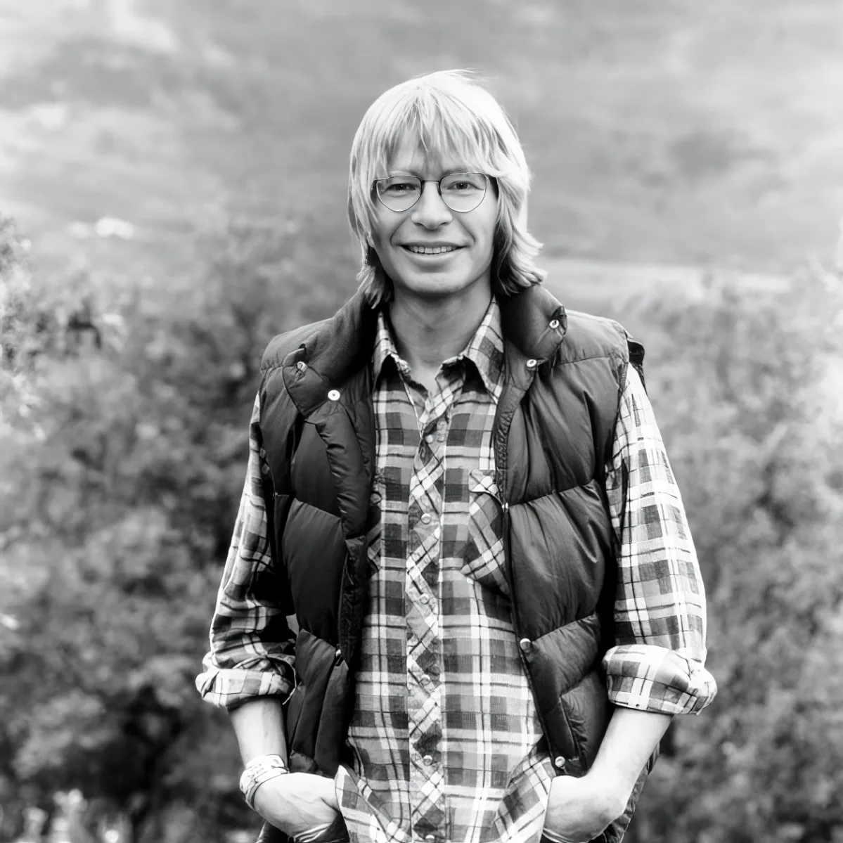 A man with blonde hair and glasses, wearing a plaid shirt and a puffer vest, stands outdoors with trees in the background.