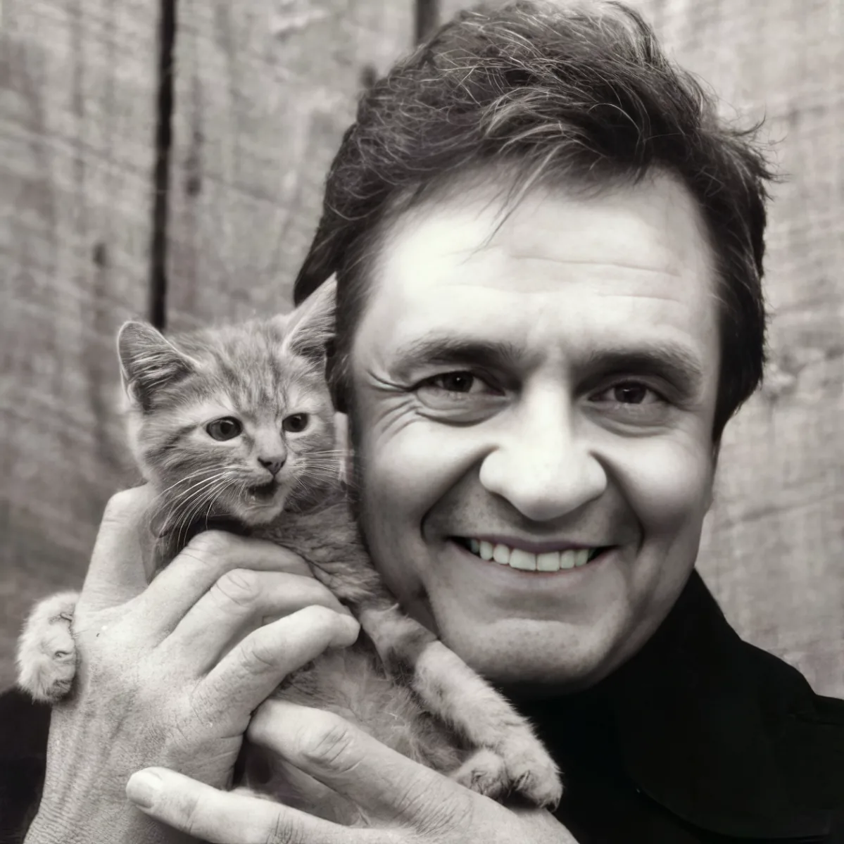 A smiling man holds a small kitten in his hands, both looking directly at the camera.