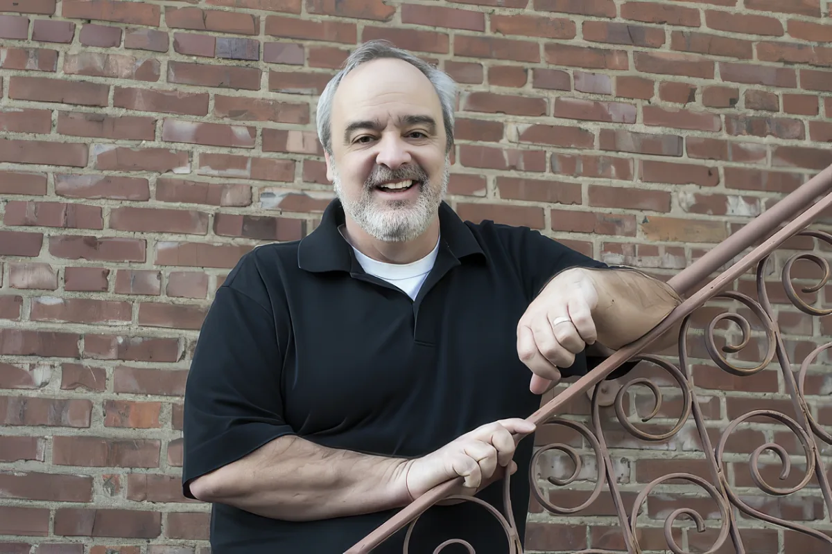 A man in a black polo shirt smiles while leaning on a ornate metal railing against a brick wall.