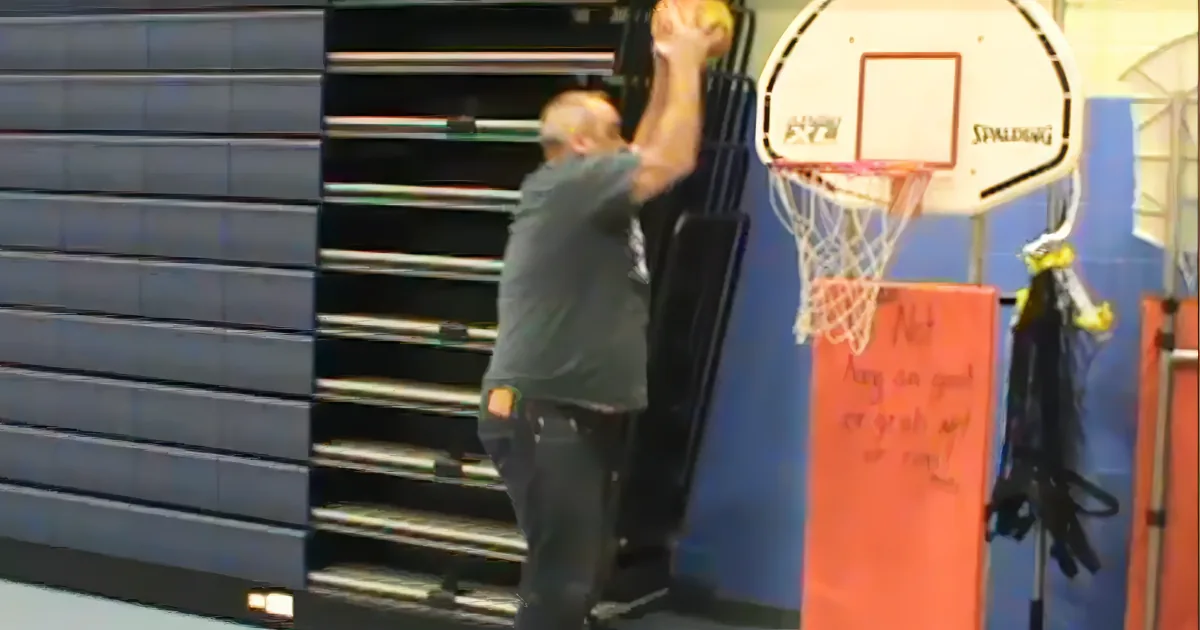 A man in a gray shirt and jeans is about to dunk a basketball into a hoop.